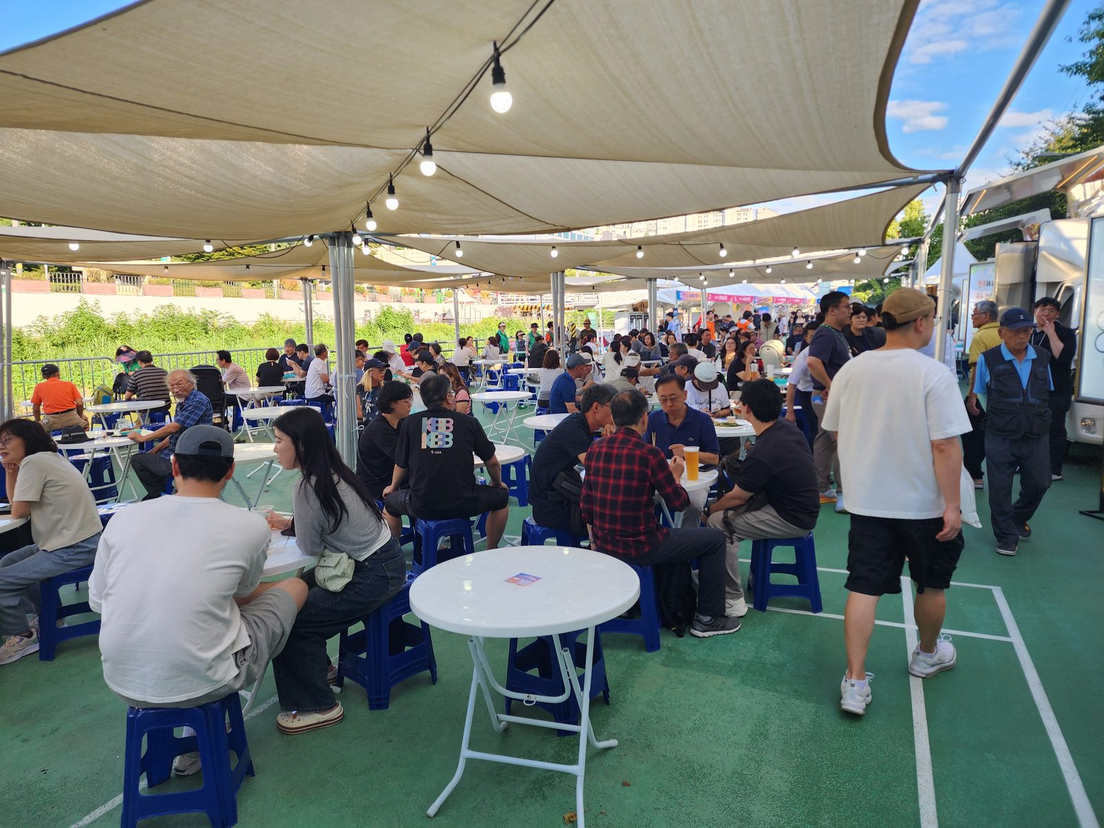 차양과 조명을 설치한 축제 취식존이 방문객으로 가득 차 있고 오른쪽 끝에 푸드트럭이 서 있다