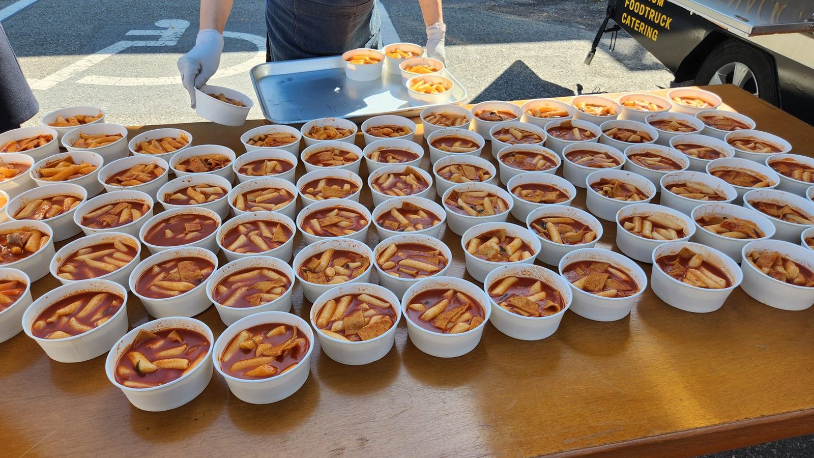 야외 배식대 위에 흰 종이 용기에 담긴 떡볶이 수십 그릇이 줄지어 놓여 있다. 붉은 국물 속에 가래떡과 어묵이 잠겨 있고, 뒤에서 장갑 낀 사람이 그릇을 쟁반에 옮기고 있으며 오른쪽 위로 푸드트럭 차체가 보인다
