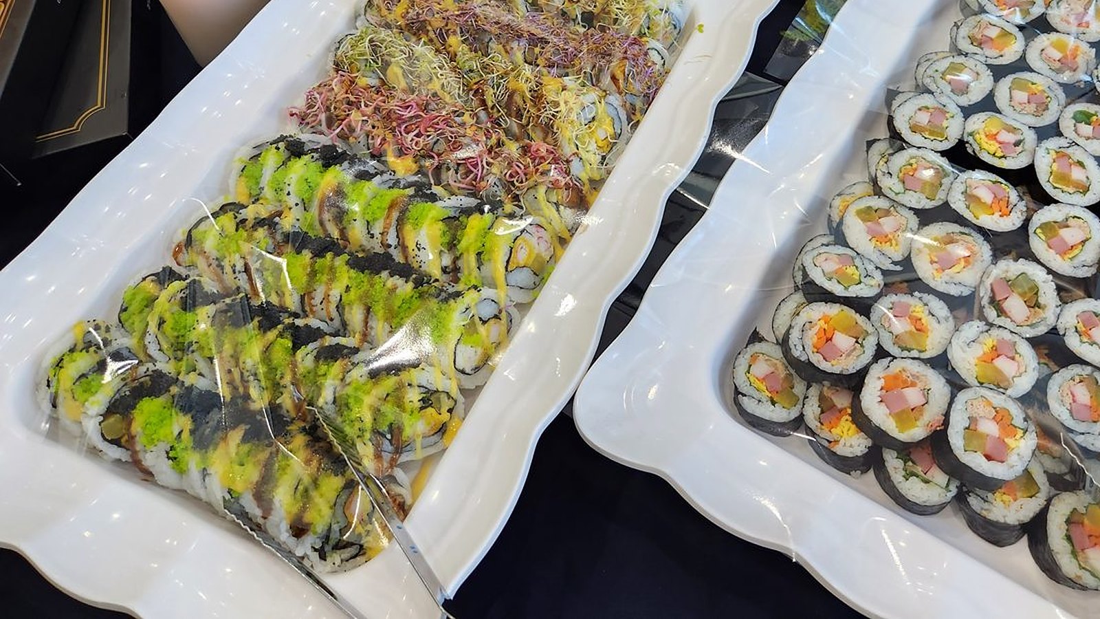 검은 식탁보 위 흰 사각 접시 두 개에 김밥이 가득 담겨 있다. 왼쪽 접시에는 밥이 바깥으로 오게 만 김밥 위에 연두색 날치알과 소스, 붉고 노란 새싹 채소를 얹은 줄들이 비스듬히 놓여 있고, 오른쪽 접시에는 햄·단무지·당근·달걀·오이가 든 둥근 김밥 단면이 빼곡하다. 접시들은 투명한 랩으로 덮여 있다