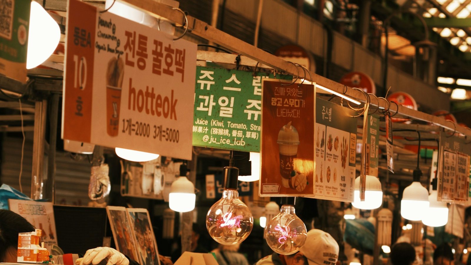 전통시장 노점 위에 「전통 꿀호떡 hotteok」이라고 쓴 간판이 조명을 받아 걸려 있고, 그 옆으로 과일 주스 간판과 전구 조명, 차양 아래 분주한 시장 통로가 이어진다