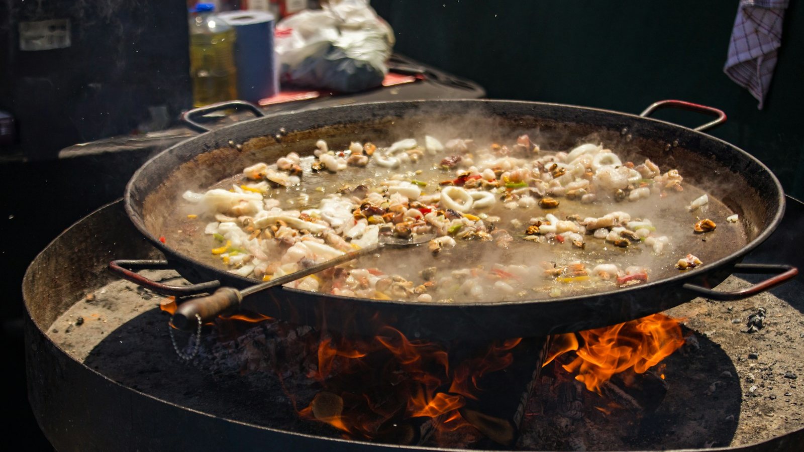 야외 화덕 위에 올린 커다란 파에야 팬에서 오징어와 홍합, 새우, 채소가 김을 내며 익고 있고, 팬 아래로 장작불이 타오르며 뒤편 작업대에는 식용유 병과 키친타월이 놓여 있다
