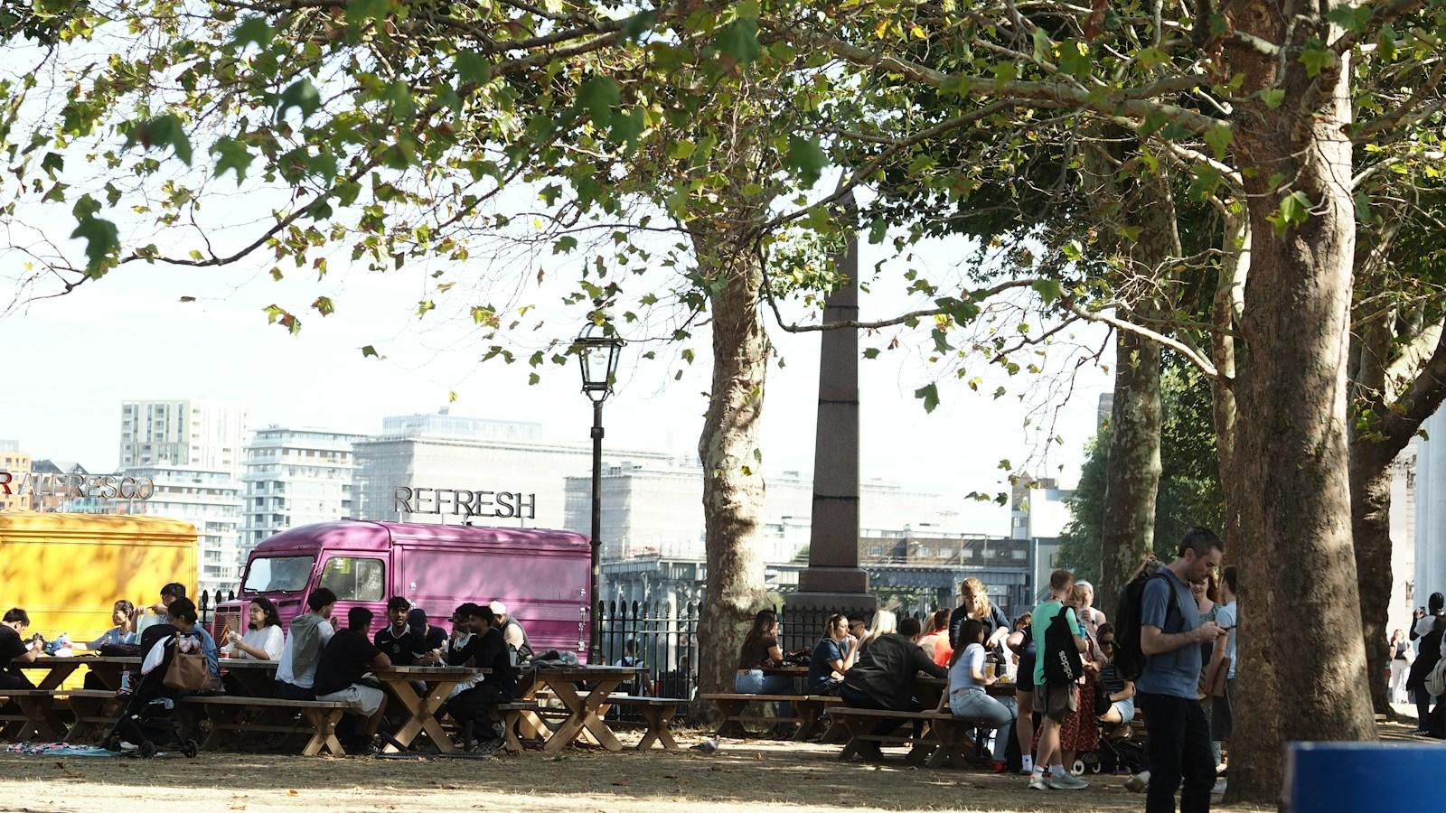 강변 공원의 큰 플라타너스 그늘 아래 나무 피크닉 테이블마다 사람들이 앉아 먹고 이야기하고 있다. 왼쪽 철제 울타리 앞에 노란색 트럭과 분홍색 클래식 밴이 서 있고, 그 너머로 강 건너편 아파트 건물들이 보인다