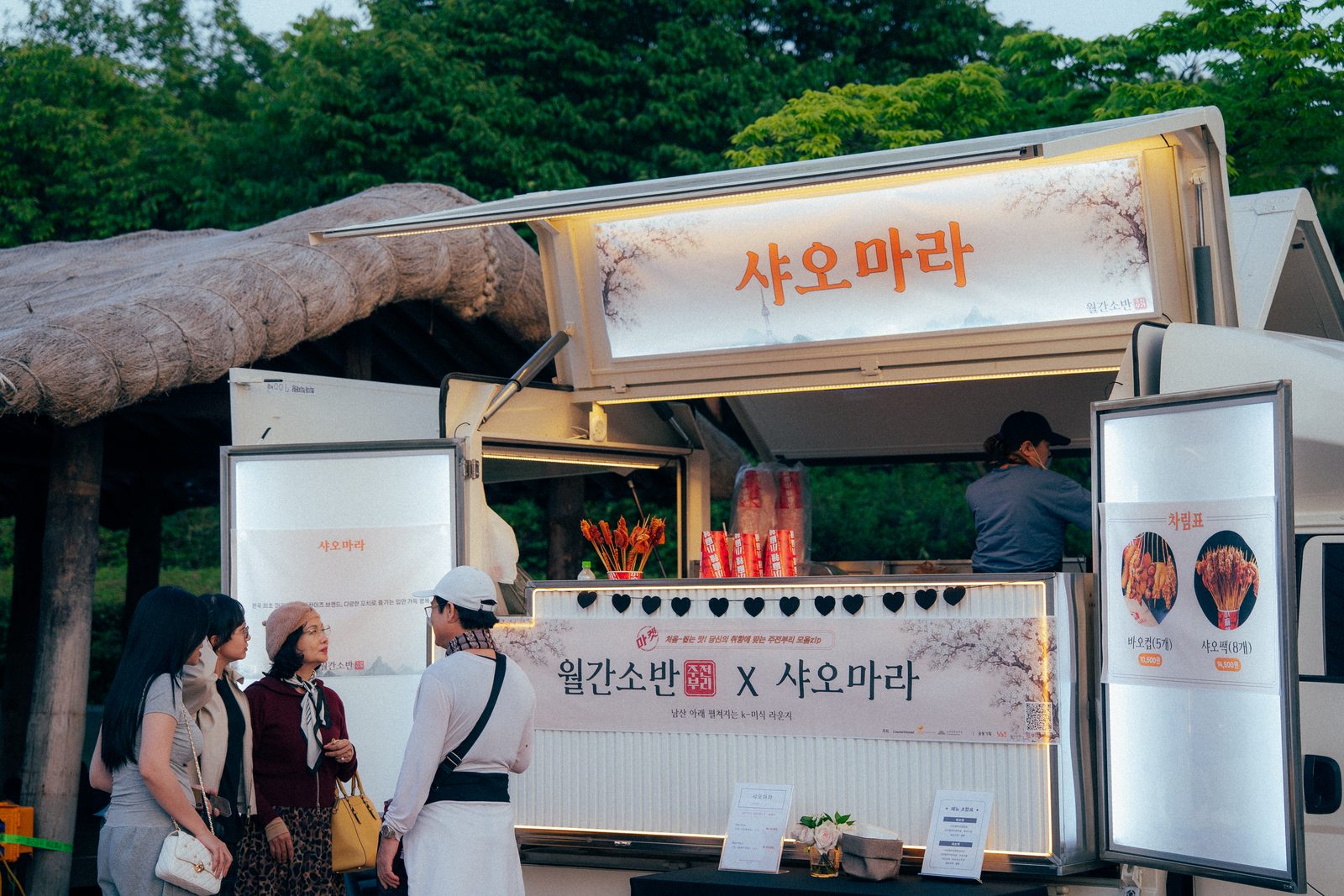 브랜드 이름을 건 조명 사이니지와 배너를 갖춘 팝업트럭 앞에서 방문객들이 메뉴를 보고 있다