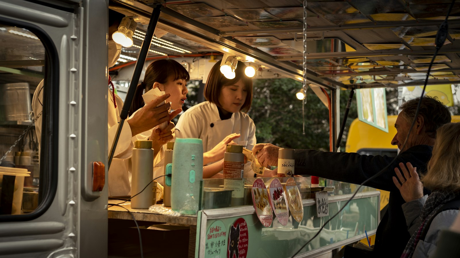조명을 켠 은색 푸드트럭 창구 안에서 흰 조리복을 입은 두 여성이 손님을 응대하고 있고, 창구 밖에서는 중년 남성이 손을 뻗어 주문한 음식을 받으며 옆에 금발의 여성이 서 있다. 창구 유리에는 일본어 메뉴 그림 카드가 붙어 있다